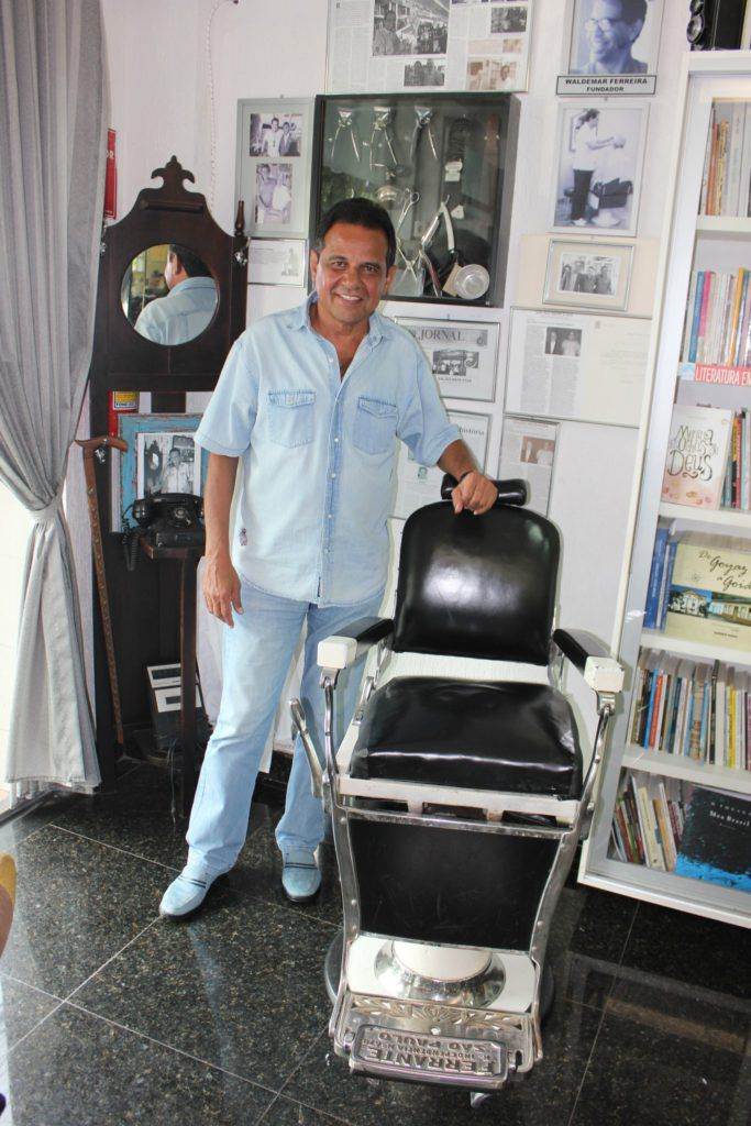 Ruimar Ferreira posa no “Cantinho Cultural” da sua barbearia, ao lado de antiga cadeira (porém conservada) herdada do pai